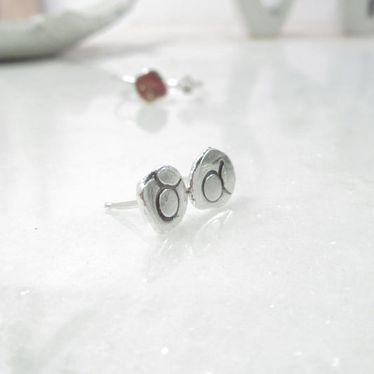 A pair of silver post earrings with oxidized astrology symbols on a white surface.