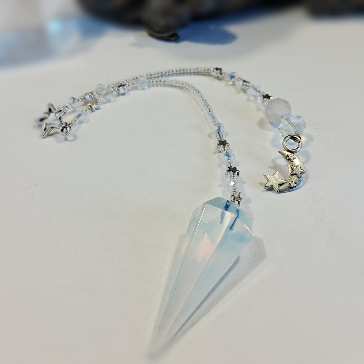 A crystal pendulum with a large faceted pale blue quartz point, suspended from a beaded chain with silver-toned star and moon charms.