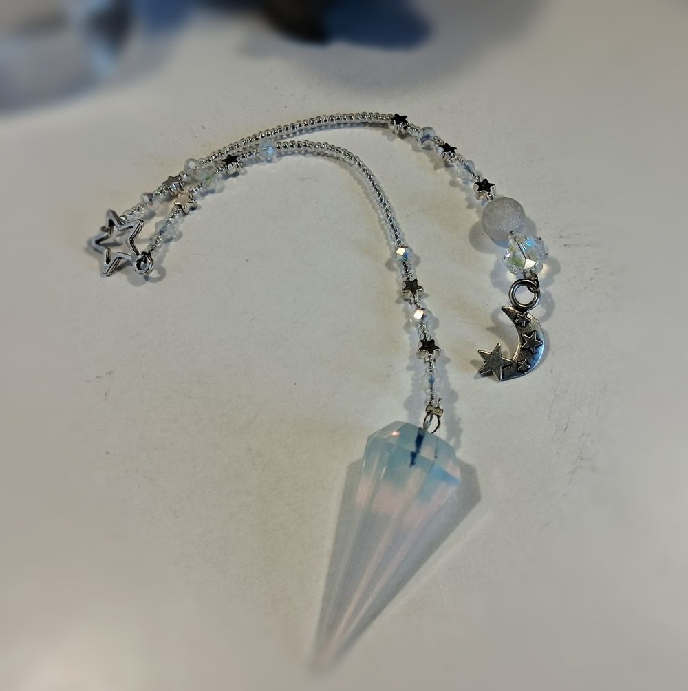 A crystal pendulum set featuring a faceted light blue and white conical crystal pendant, suspended from a beaded chain with silver-toned charms including a star, moon, and small beads.