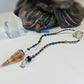 A beaded pendulum with a cone-shaped crystal pendant and a silver-toned tree of life clasp, displayed on a white surface with a blurred statue and clear crystal in the background.