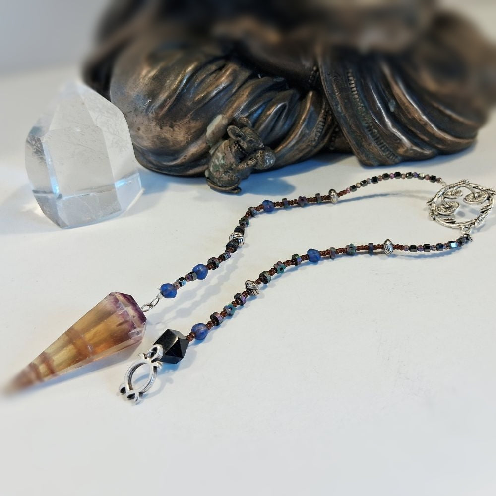 A beaded pendulum with a cone-shaped crystal pendant and a silver-toned tree of life clasp, displayed on a white surface with a blurred statue and clear crystal in the background.