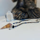 A beaded spiritual Flurite Pendulum with a brown crystal cone pendant and silver-toned charms, displayed on a white surface next to a clear crystal point and a blurred dark figurine.