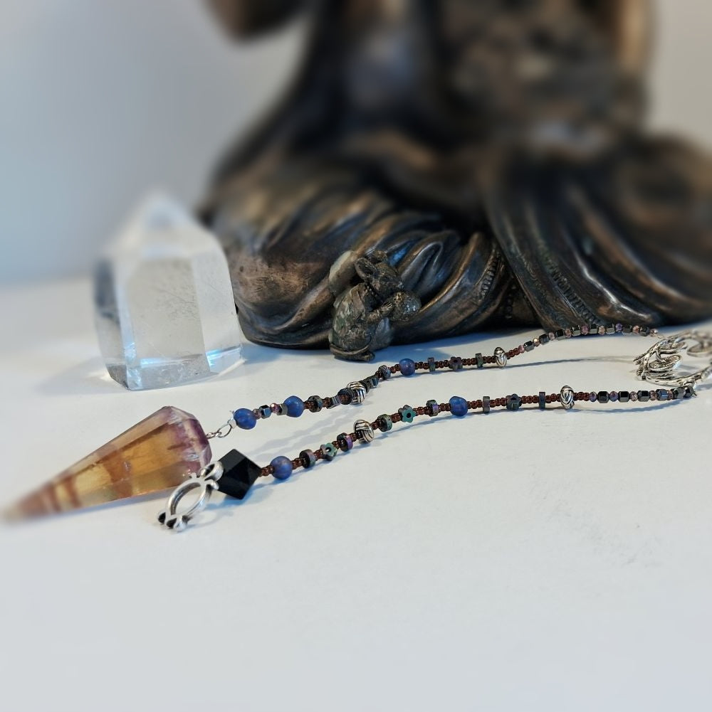 A beaded spiritual Flurite Pendulum with a brown crystal cone pendant and silver-toned charms, displayed on a white surface next to a clear crystal point and a blurred dark figurine.