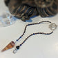 A beaded magic mushroom pendulum with a pointed crystal pendant, featuring dark brown and blue beads, silver-toned metal spacers, a silver spiral clasp, and a small black bead, displayed on a white surface with a blurred decorative object in the background.