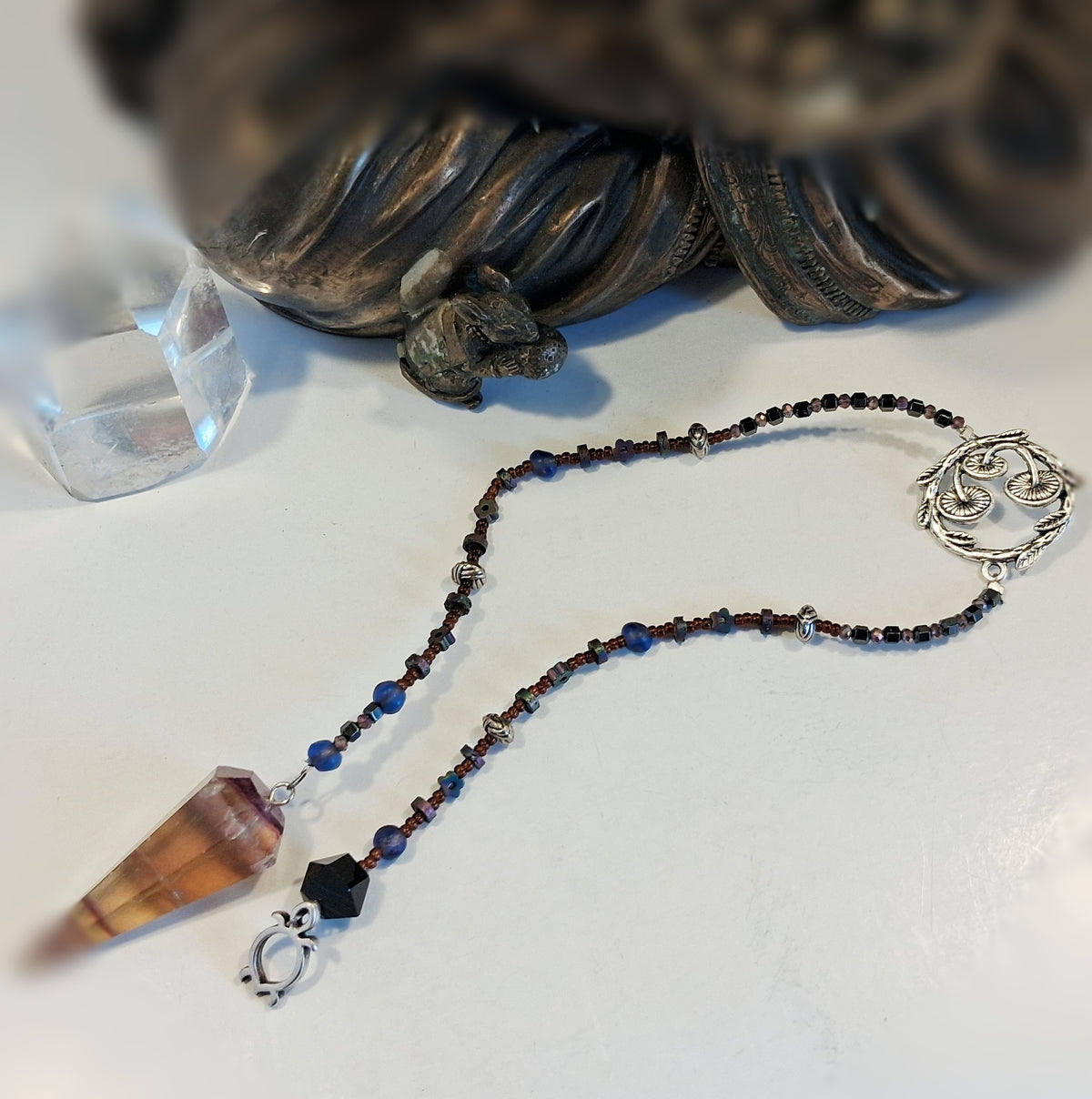 A beaded magic mushroom pendulum with a pointed crystal pendant, featuring dark brown and blue beads, silver-toned metal spacers, a silver spiral clasp, and a small black bead, displayed on a white surface with a blurred decorative object in the background.