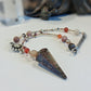 A beaded spiritual pendulum featuring a large pointed crystal cone pendant, red agate beads, and silver-toned charms, displayed on a white surface.