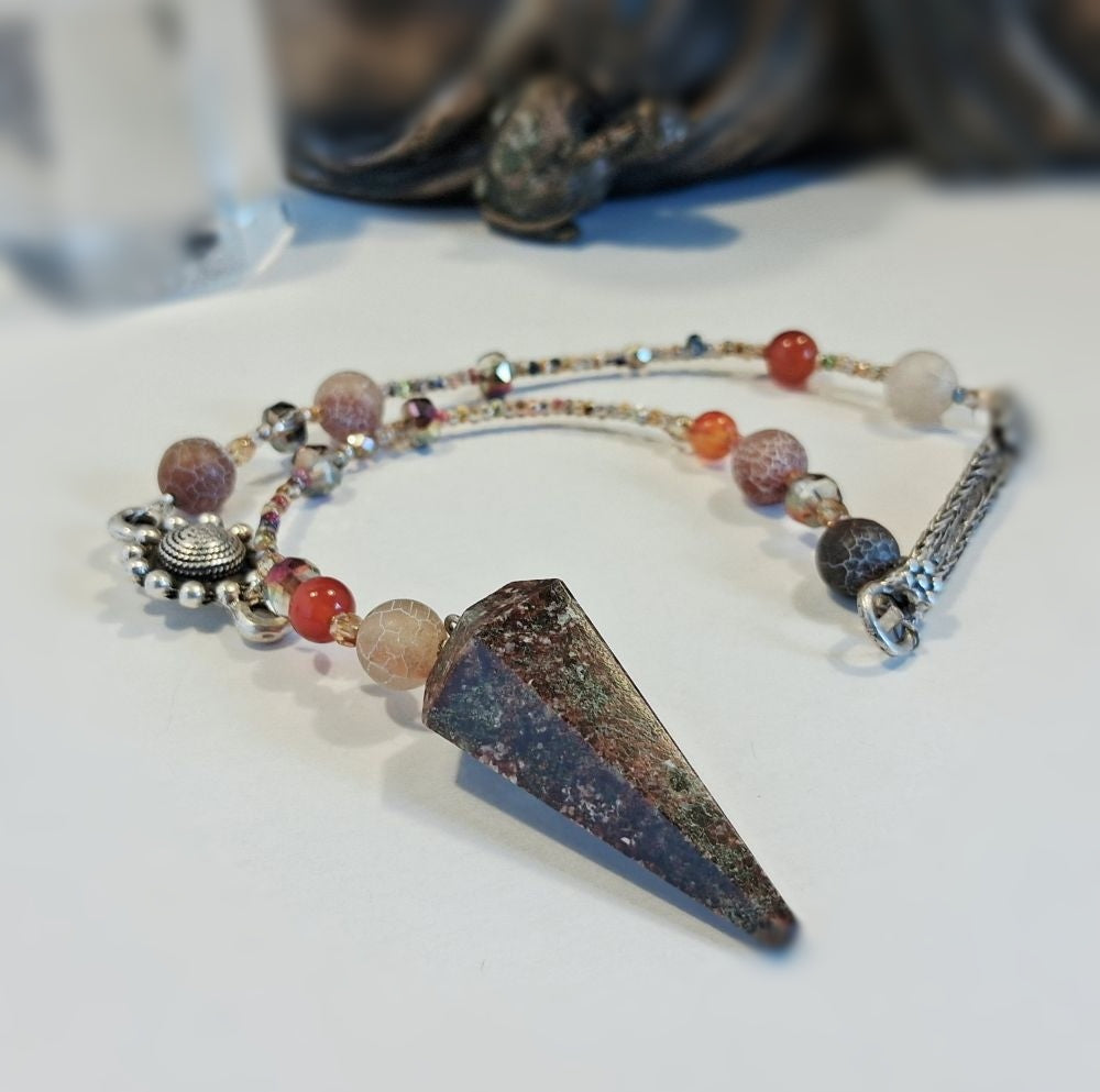 A beaded spiritual pendulum featuring a large pointed crystal cone pendant, red agate beads, and silver-toned charms, displayed on a white surface.