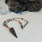 A beaded spiritual pendulum featuring a prominent garnet crystal, silver-toned metal charms, and multi-colored beads, displayed on a white surface with a blurred dark figurine in the background.