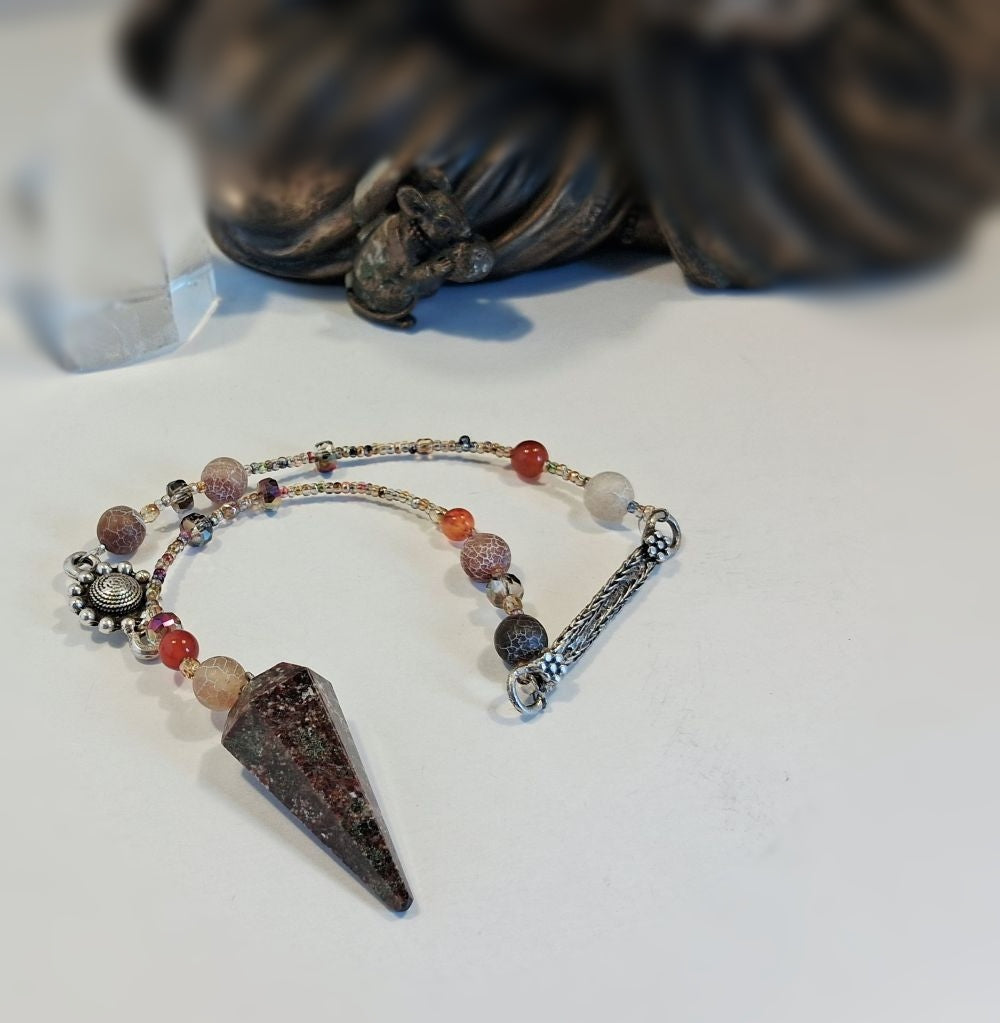 A beaded spiritual pendulum featuring a prominent garnet crystal, silver-toned metal charms, and multi-colored beads, displayed on a white surface with a blurred dark figurine in the background.