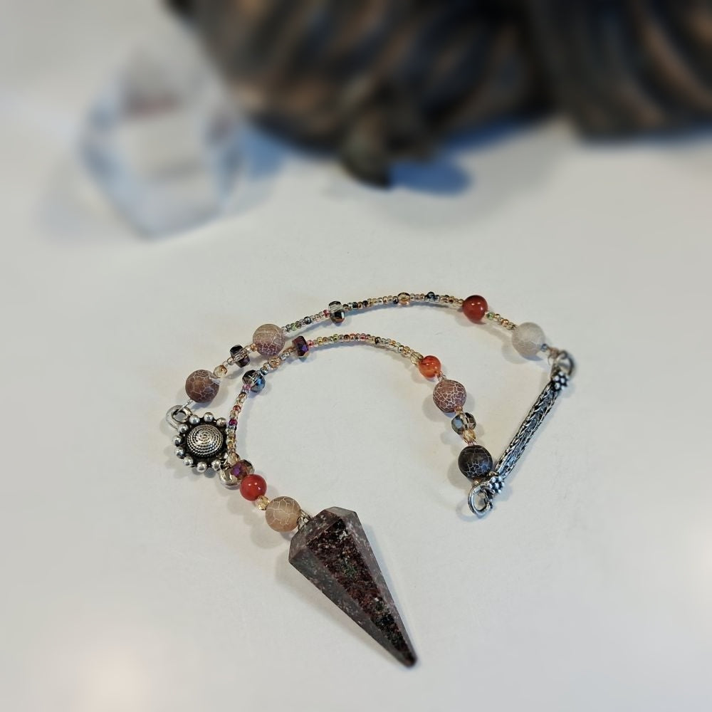 A beaded crystal pendulum featuring a large garnet crystal pendant, small red beads, and silver-toned metal charms, displayed on a white surface.