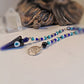 A beaded evil eye pendulum with a large blue glass cone charm, a round silver-tone metal pendant engraved with an eye of providence, and blue, turquoise, and black beads, displayed on a light surface with a blurred statue in the background.