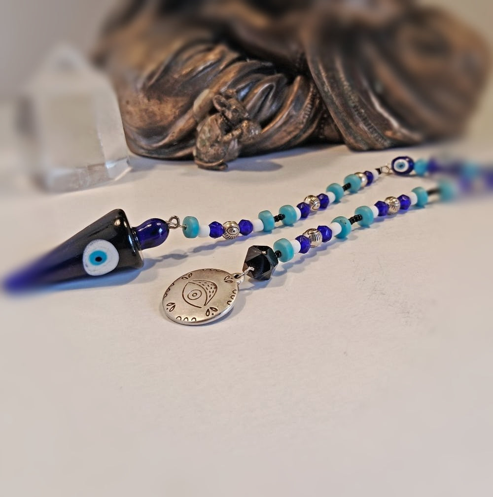 A beaded evil eye pendulum with a large blue glass cone charm, a round silver-tone metal pendant engraved with an eye of providence, and blue, turquoise, and black beads, displayed on a light surface with a blurred statue in the background.