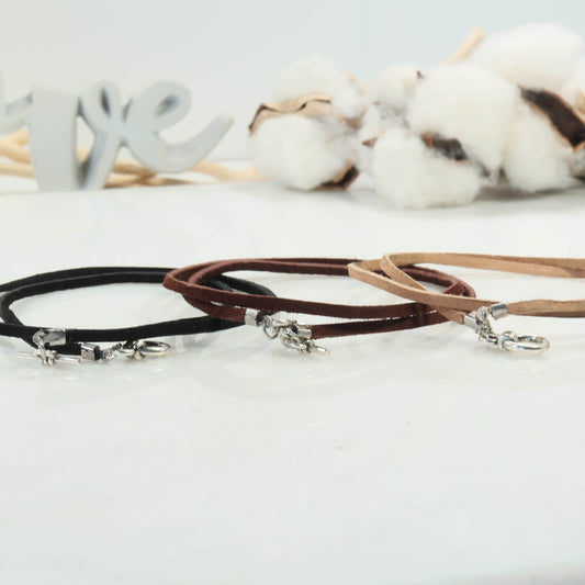 Three suede necklaces in black, brown, and beige on a light background with cotton and wooden elements.
